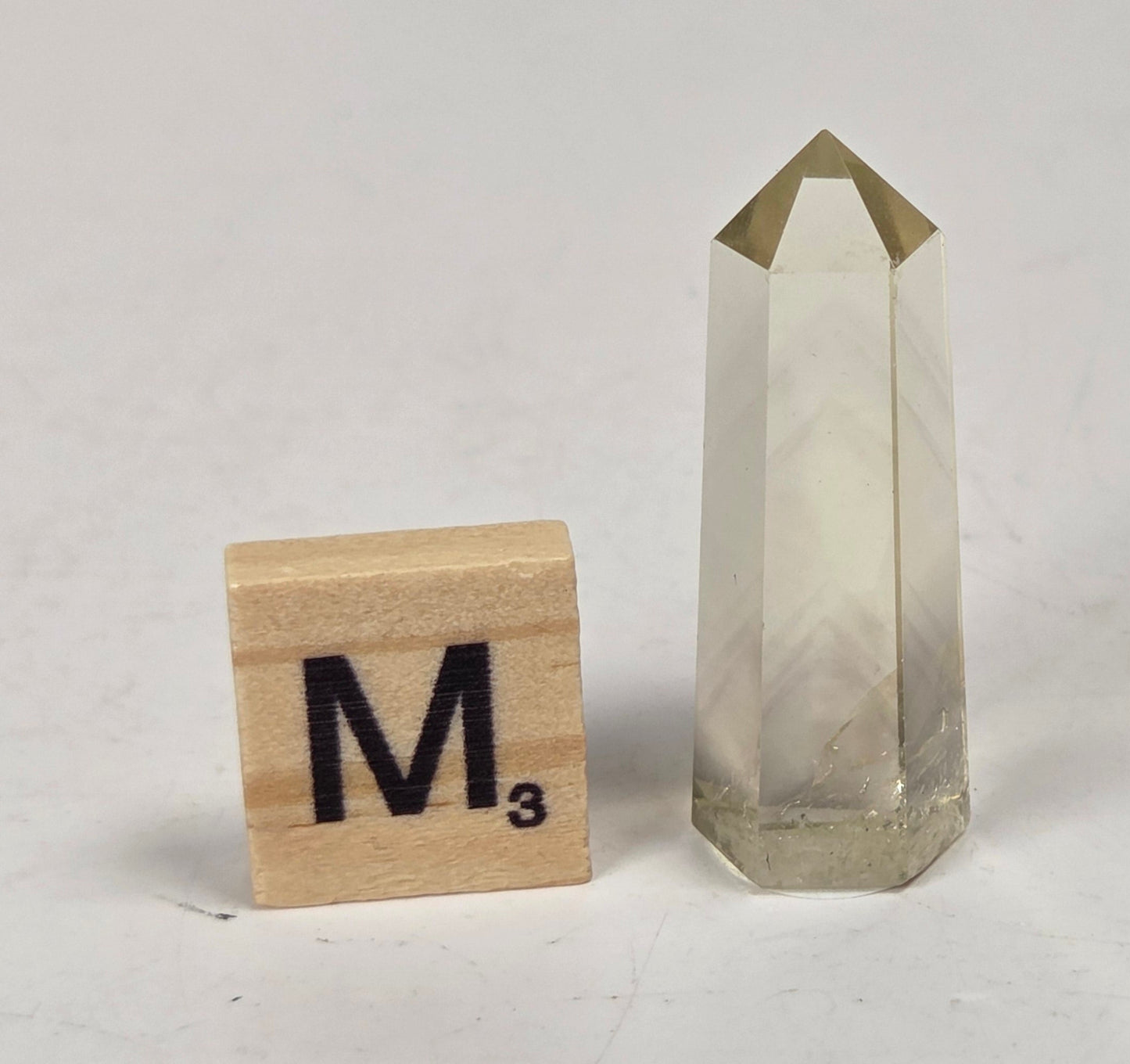 A clear faceted crystal point tower with a gold-toned pyramid cap, shown on a light marble-like surface next to a wooden Scrabble tile marked M³.