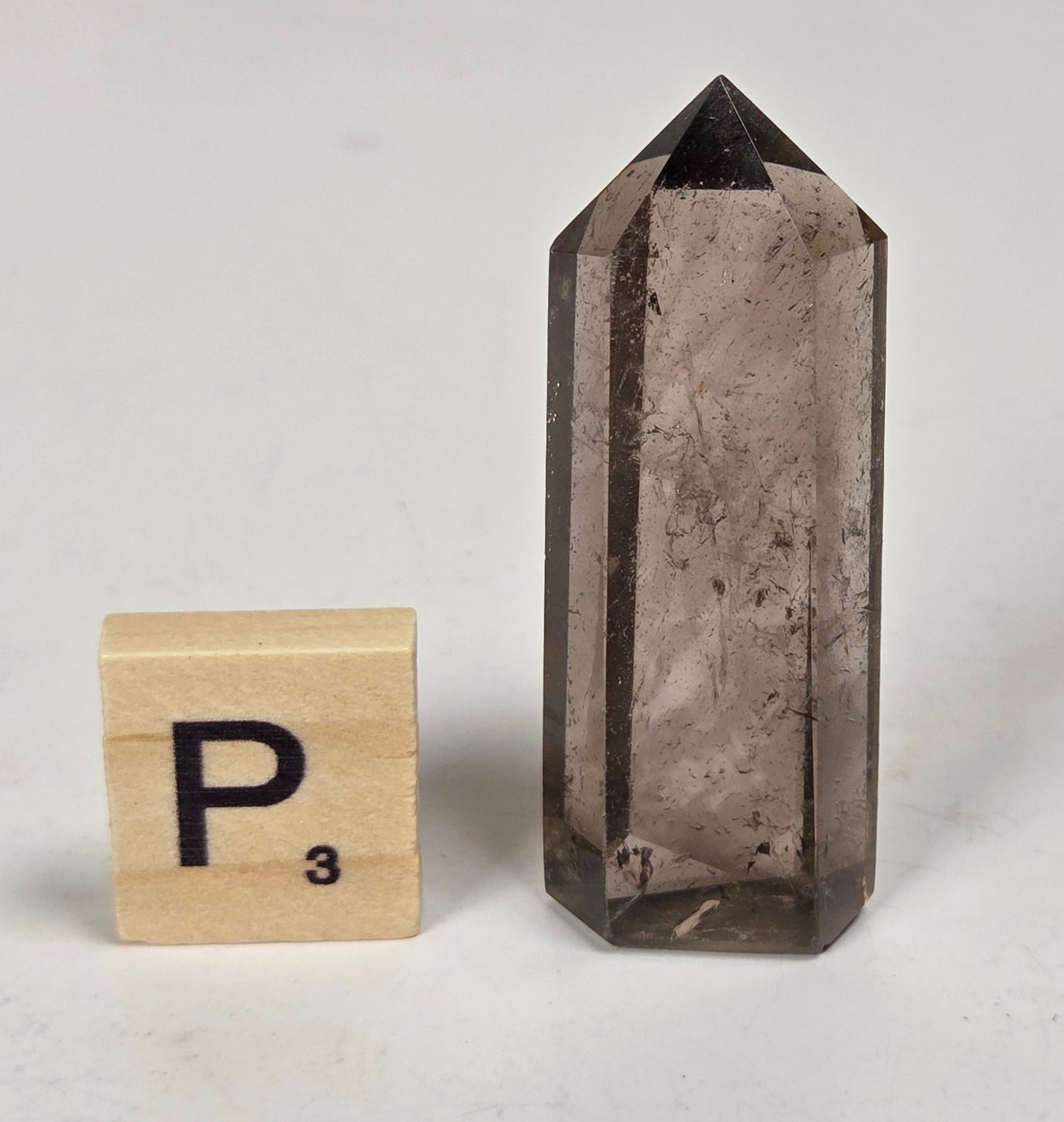 A smoky quartz crystal point standing upright next to a wooden Scrabble tile showing the letter P, on a light gray surface.