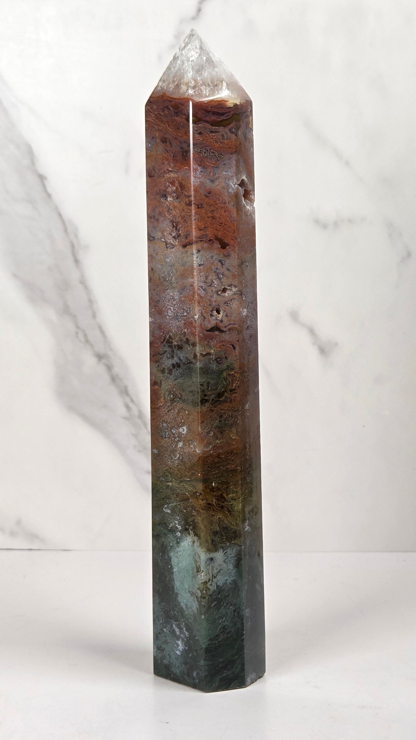 A polished natural crystal tower with a pointed apex, featuring layered earthy colors including brown, green, and dark gray, set against a white marble background.