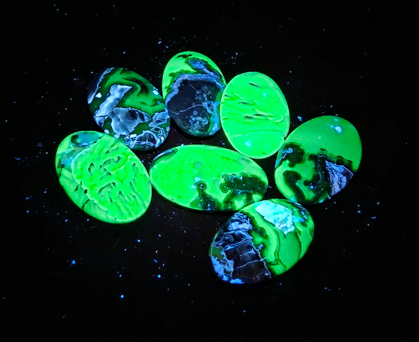 Seven oval cabochon gemstones with a glowing neon green and black marbled pattern, arranged on a solid black background under UV light.