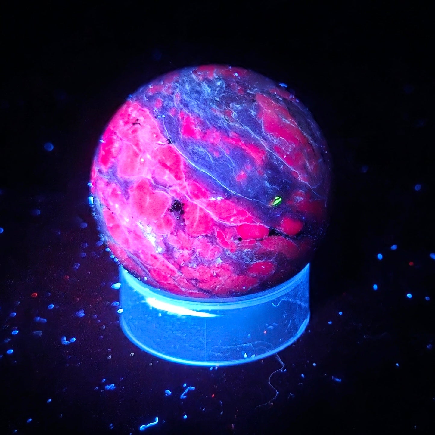 A glowing marbled crystal ball with vibrant pink, purple, and blue fluorescence, displayed on a matching blue cylindrical stand against a black background.