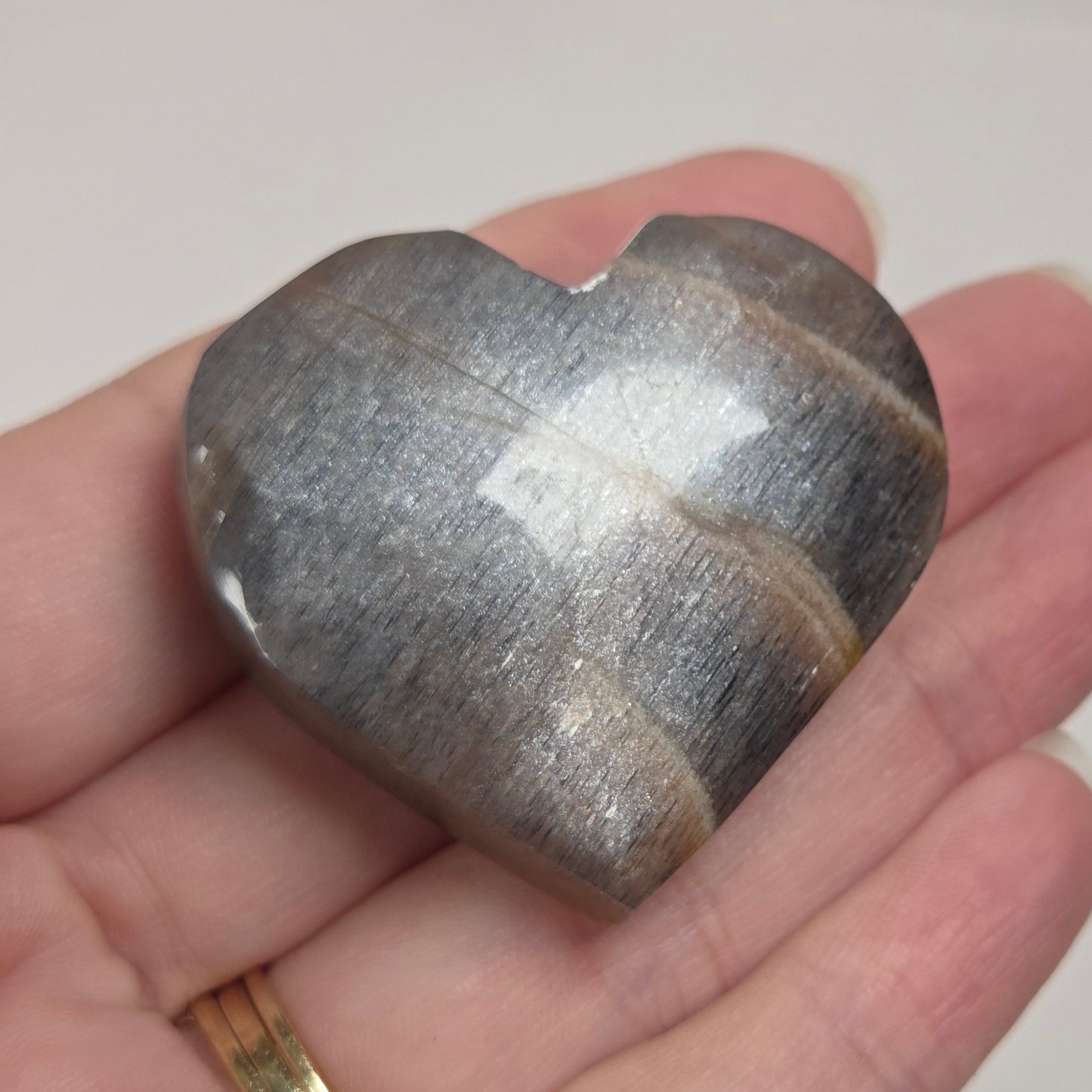 A polished heart-shaped stone with natural banding, held in a person's hand. The stone has a metallic silver-gray appearance with darker brownish-gray bands, and the hand is wearing a gold ring.