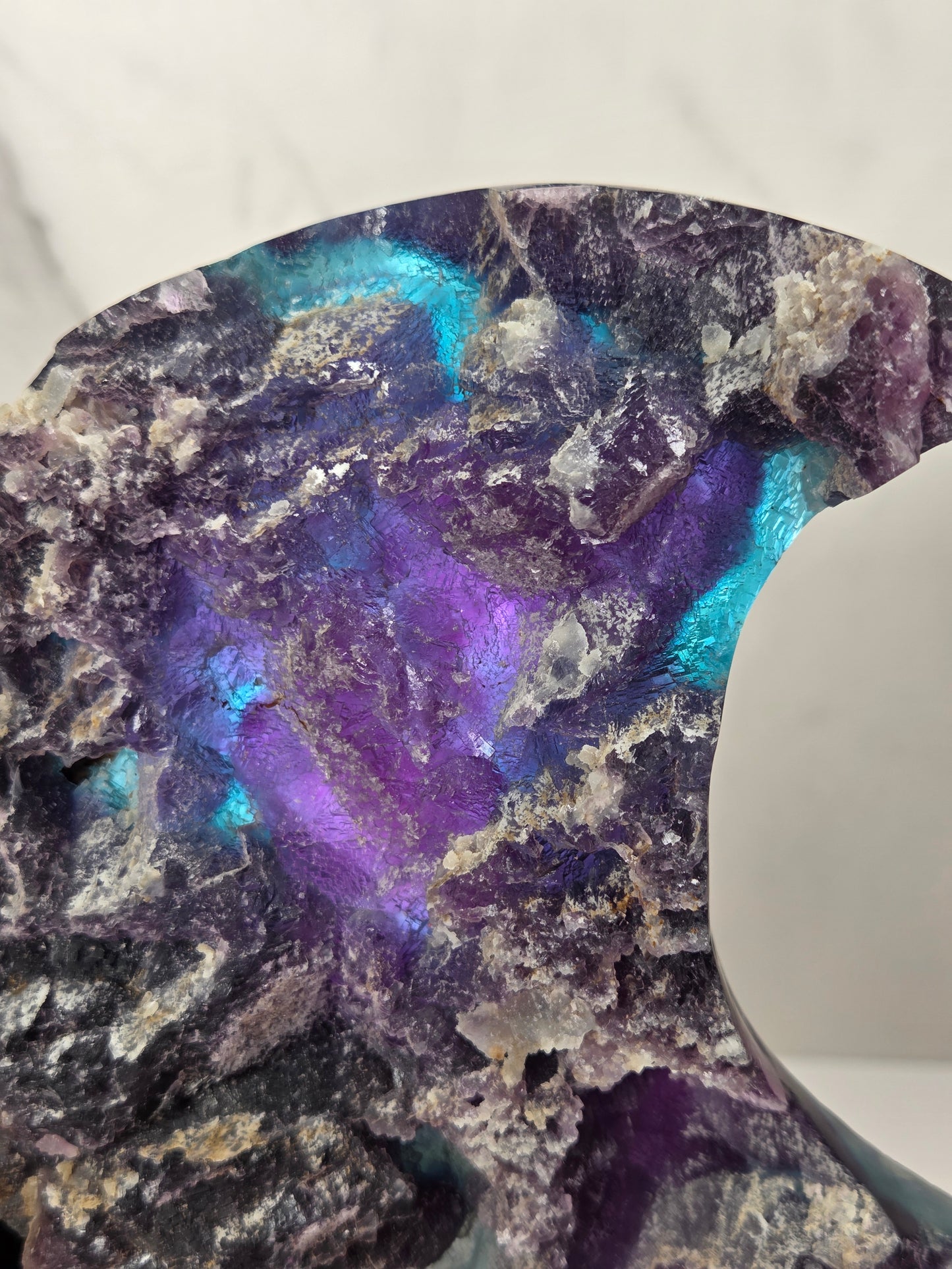 A close-up of a rough, polished stone or mineral specimen with a highly iridescent surface showing vivid purple, blue, and teal shifts over a dark base, photographed against a light marble-like background.