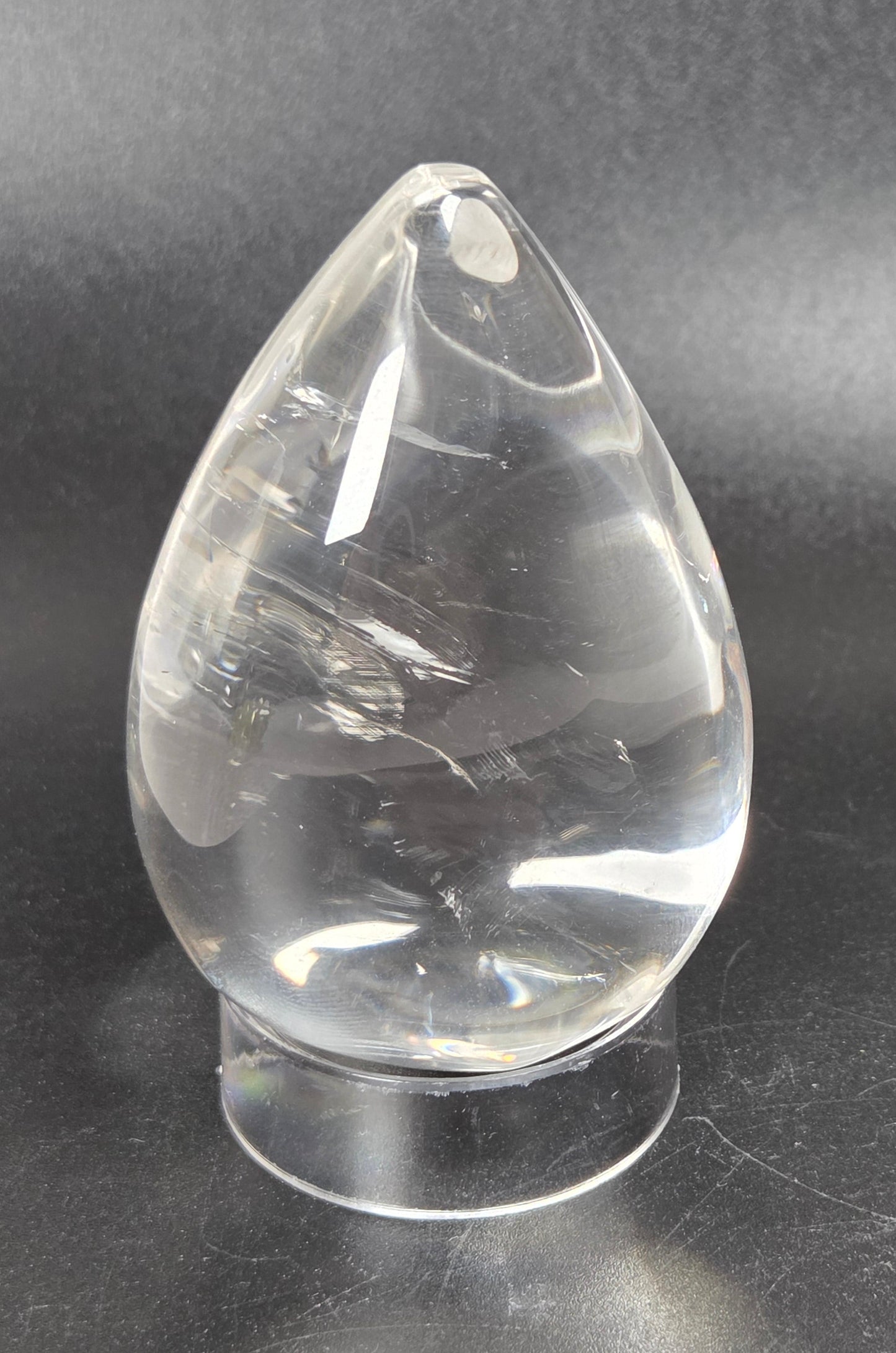 A clear teardrop-shaped glass or crystal object with a rounded base, photographed on a dark gray to black background.