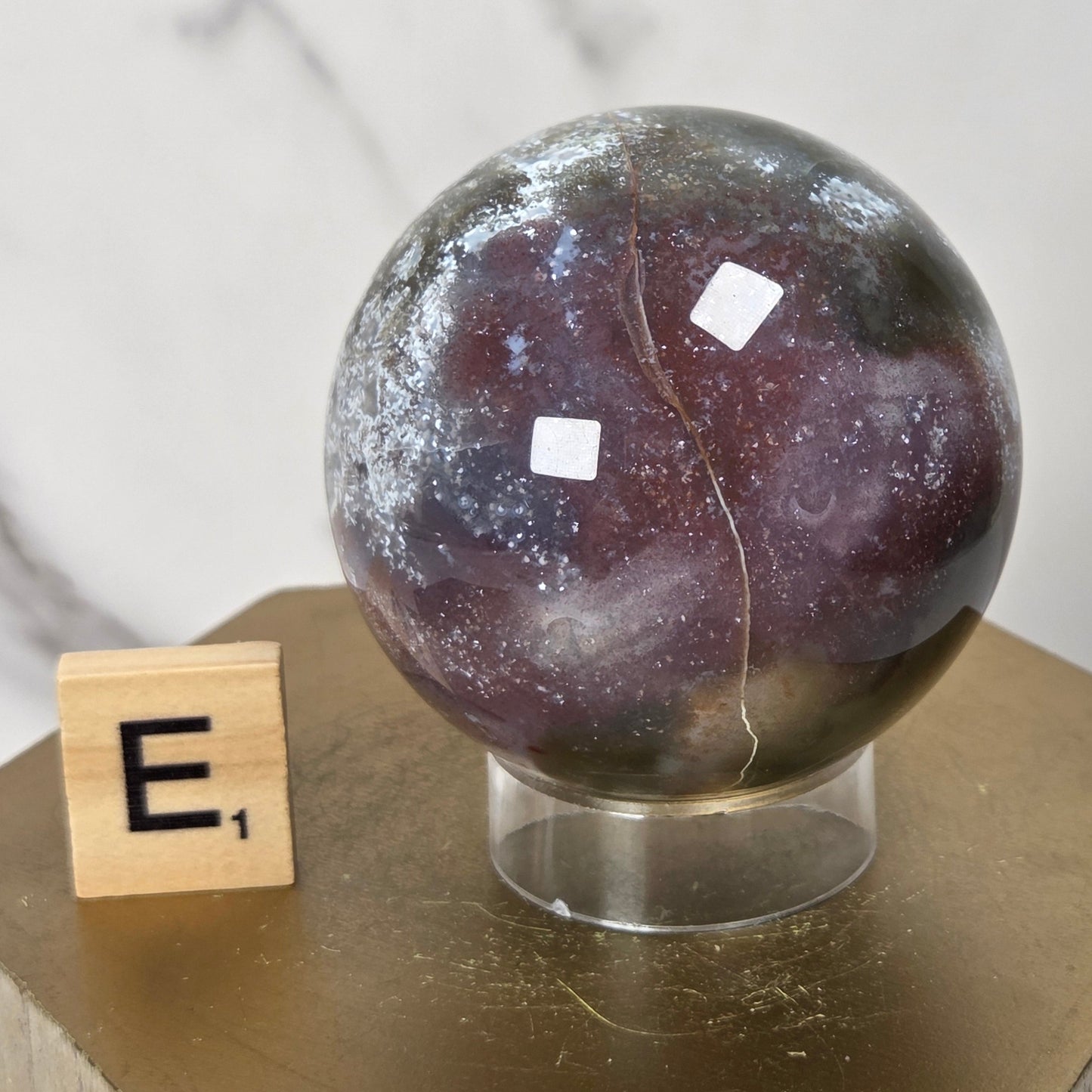A polished crystal sphere with a marbled pattern of purple, gray, and white, displayed on a clear stand next to a wooden Scrabble tile on a gold hexagonal pedestal.