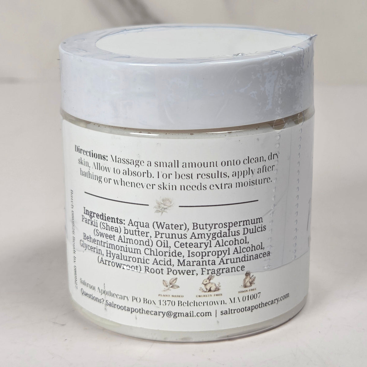 A white cosmetic jar with a white lid and a white label showing the back side with product ingredients and directions, set against a light marble background.