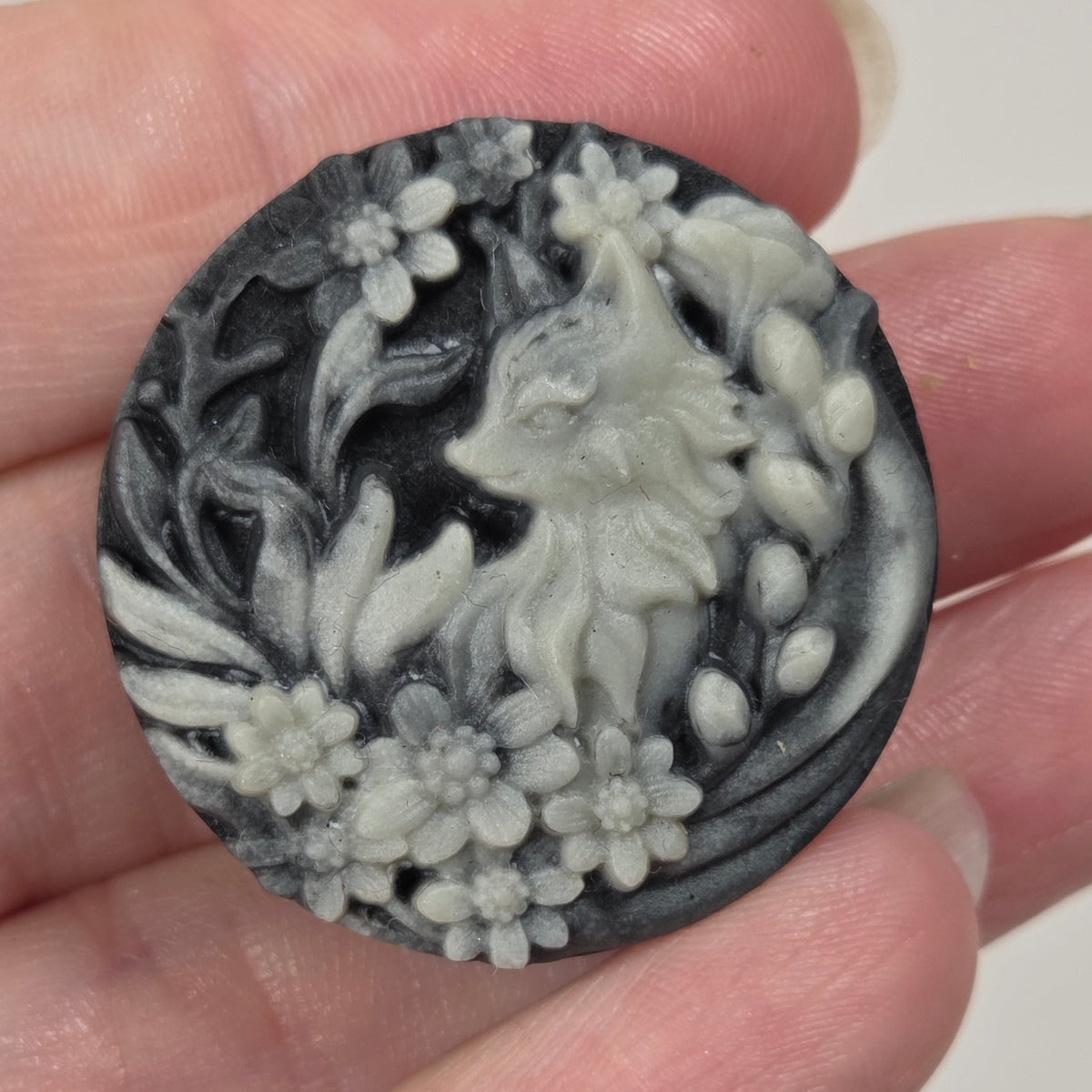 A round carved stone or resin cabochon featuring a fox surrounded by flowers, held in a person's hand. The cabochon has a dark charcoal and light gray marbled pattern.