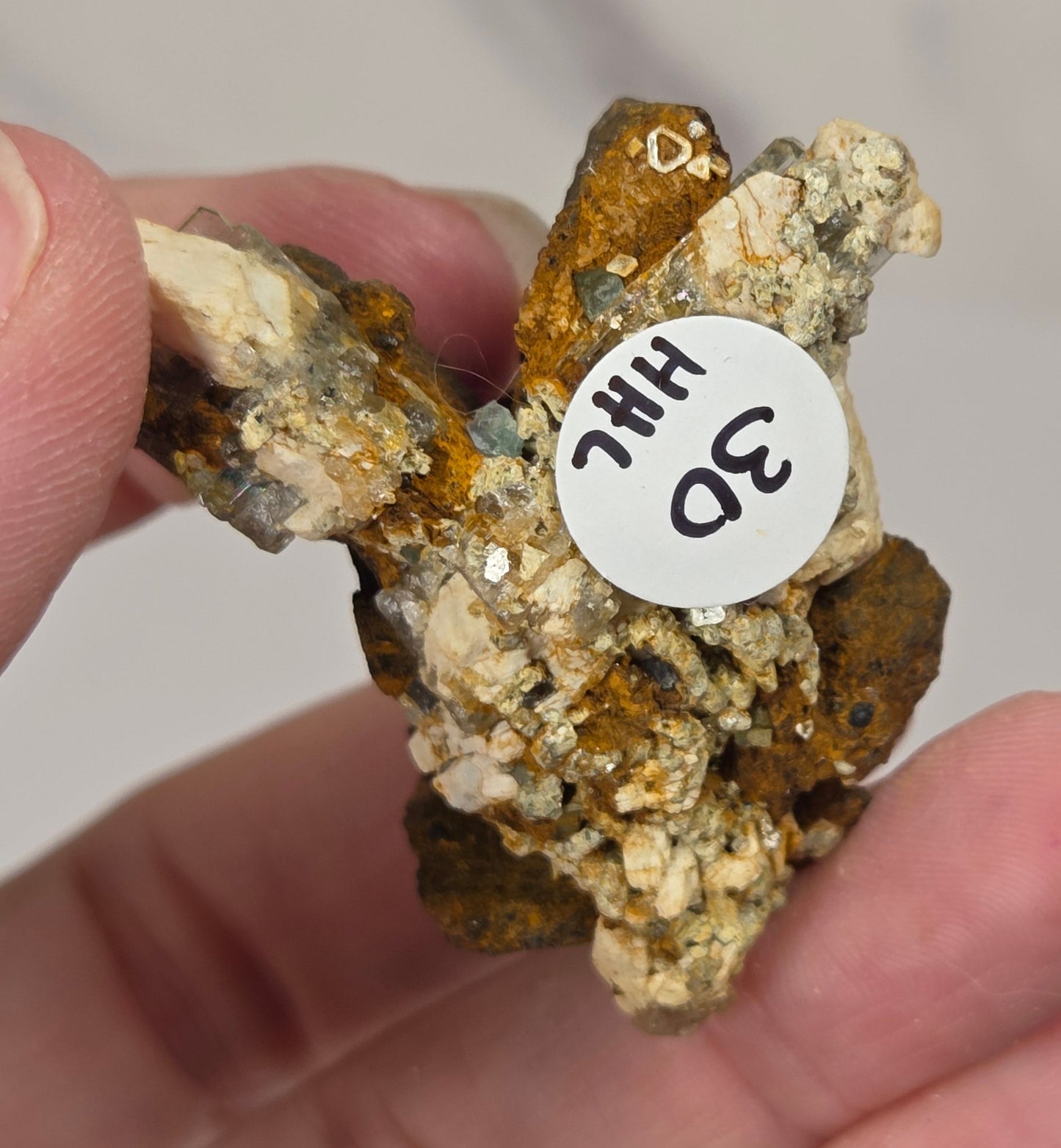 A hand holding a small mineral specimen with a white circular tag that reads '3D HHC'. The specimen features a mix of golden-brown, orange, and off-white crystalline textures.