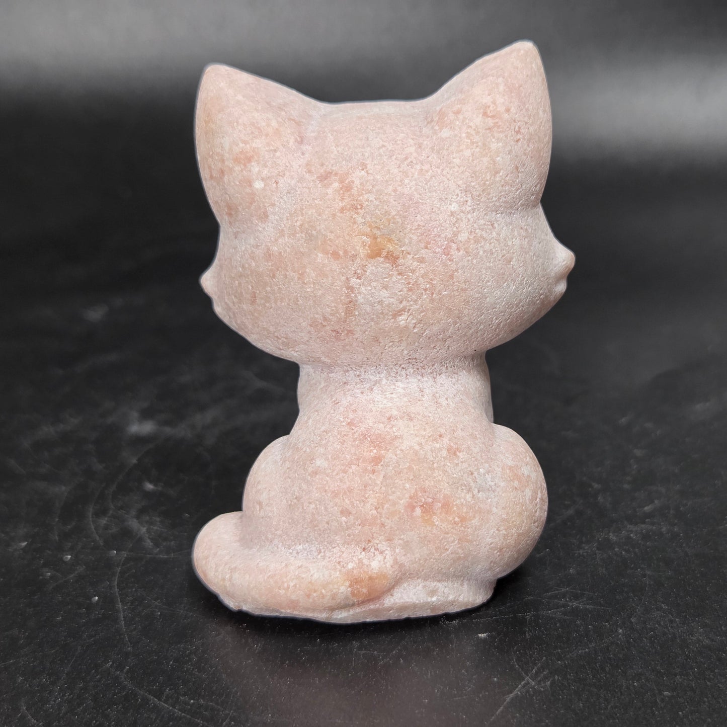 A small pink cat figurine shown from the back, made of a textured stone-like material, set against a dark gray background.