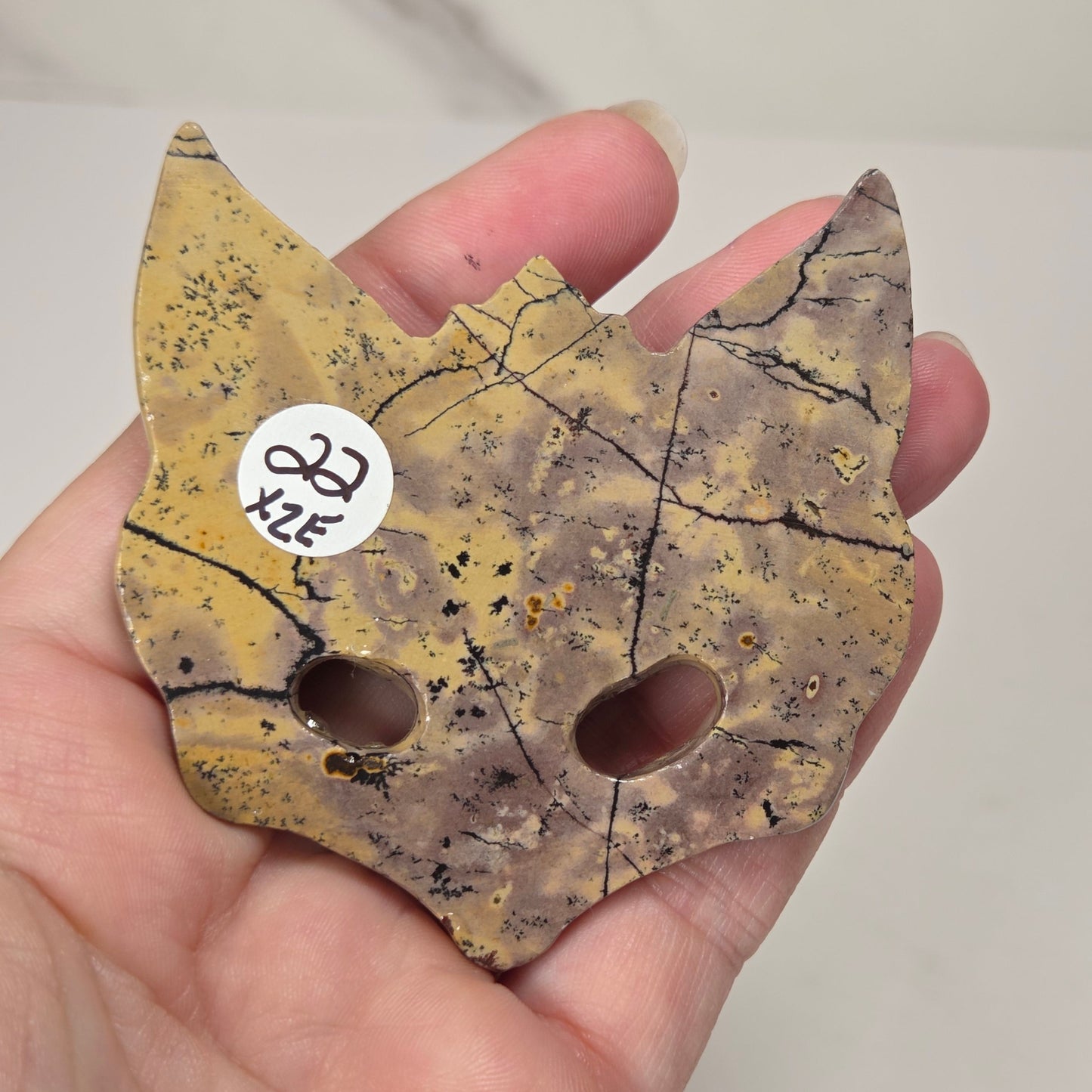 A hand holding a small cat-shaped decorative piece made from a mottled yellow and brown stone with black veining and two circular cutouts. A small white sticker with handwritten text is attached near one ear.