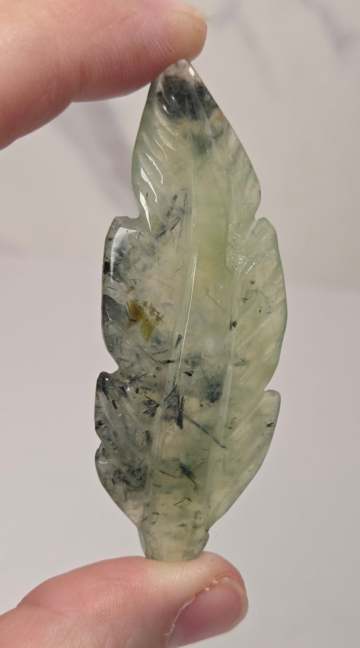 A hand holding a carved feather-shaped pendant made of milky translucent pale green stone with dark inclusions, shown against a soft light gray background.