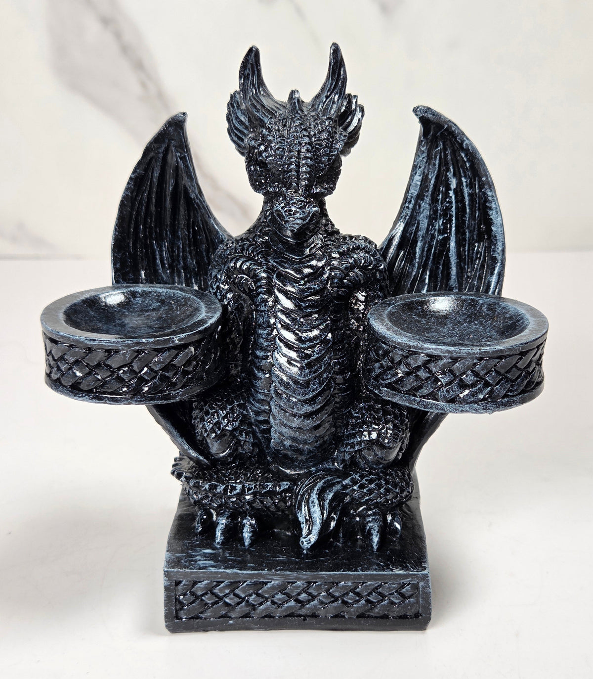 A black dragon figurine candle holder with outstretched wings and two cup-like holders on the sides, shown against a light marble-patterned background.