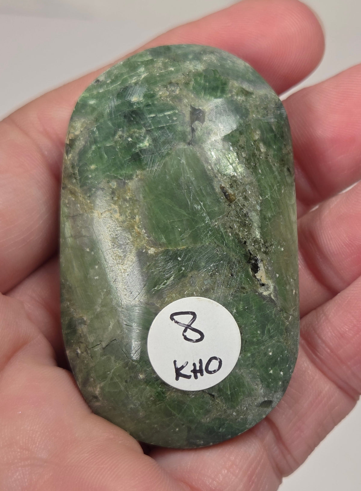 Hand holding a polished green diopside gemstones with a bottle green, brownish green or light green color, displayed on a white surface.