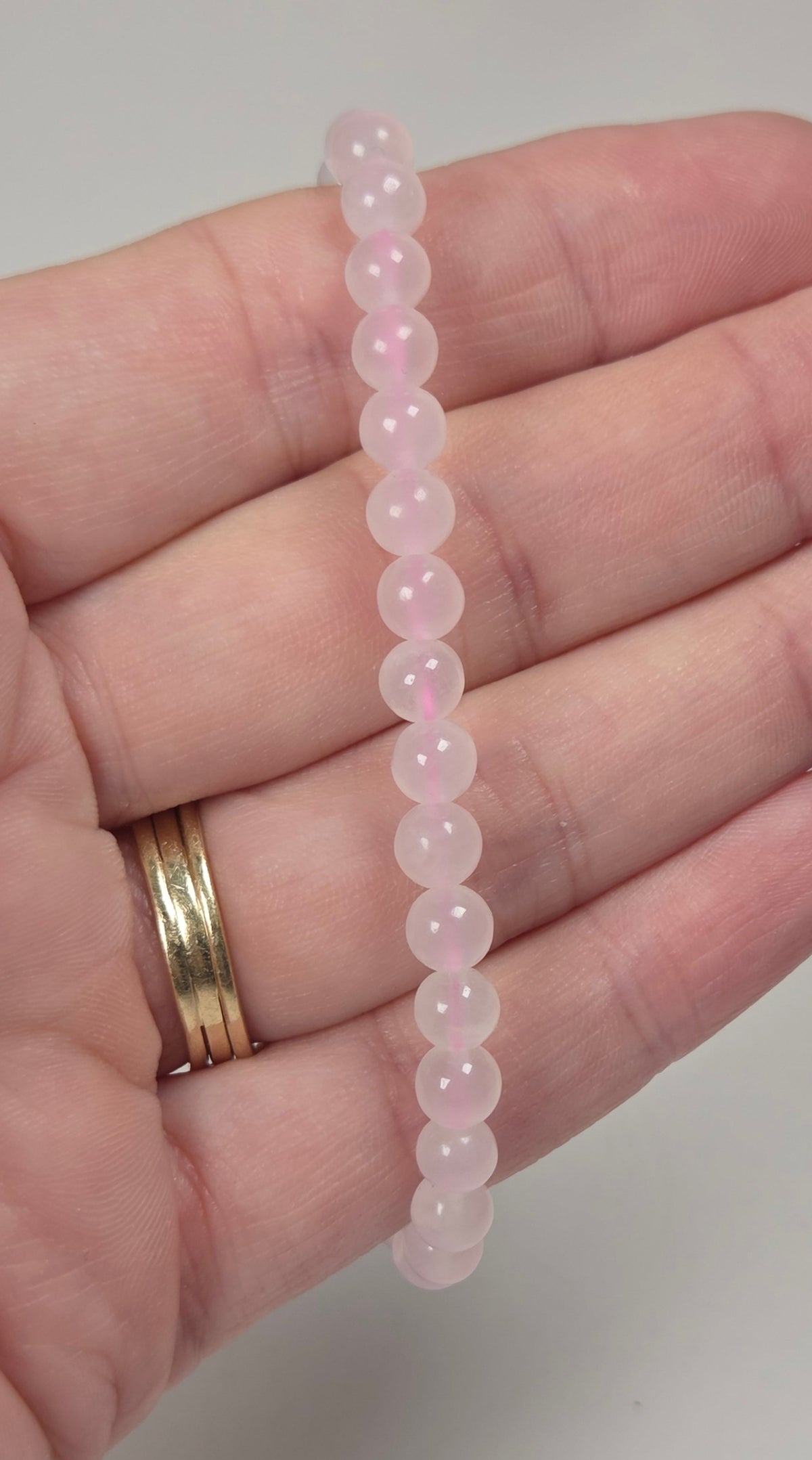 A pink beaded stretch bracelet made of smooth, round translucent beads, held in a person's hand next to a gold ring.