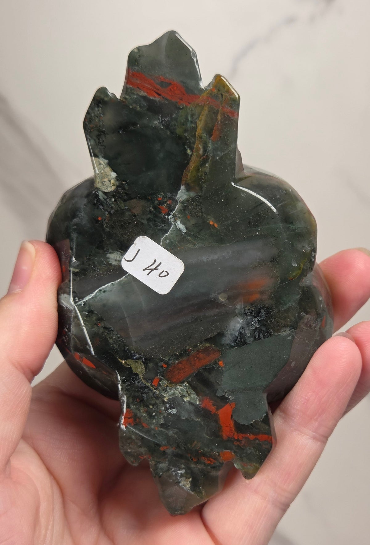 A carved stone rose sculpture with red, brown, and gray marbled petals on a silver metal stem and round base, displayed on a white surface with a small wooden letter tile nearby.