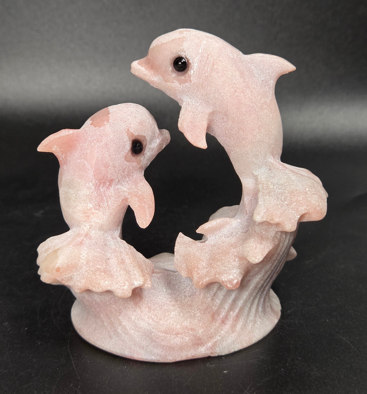 A pale pink carved figurine of two dolphins rising from stylized waves on a dark black background.