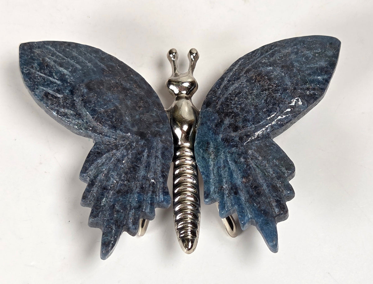 A butterfly-shaped brooch featuring textured dark blue-gray wings and a metallic silver body, displayed on a light off-white background.