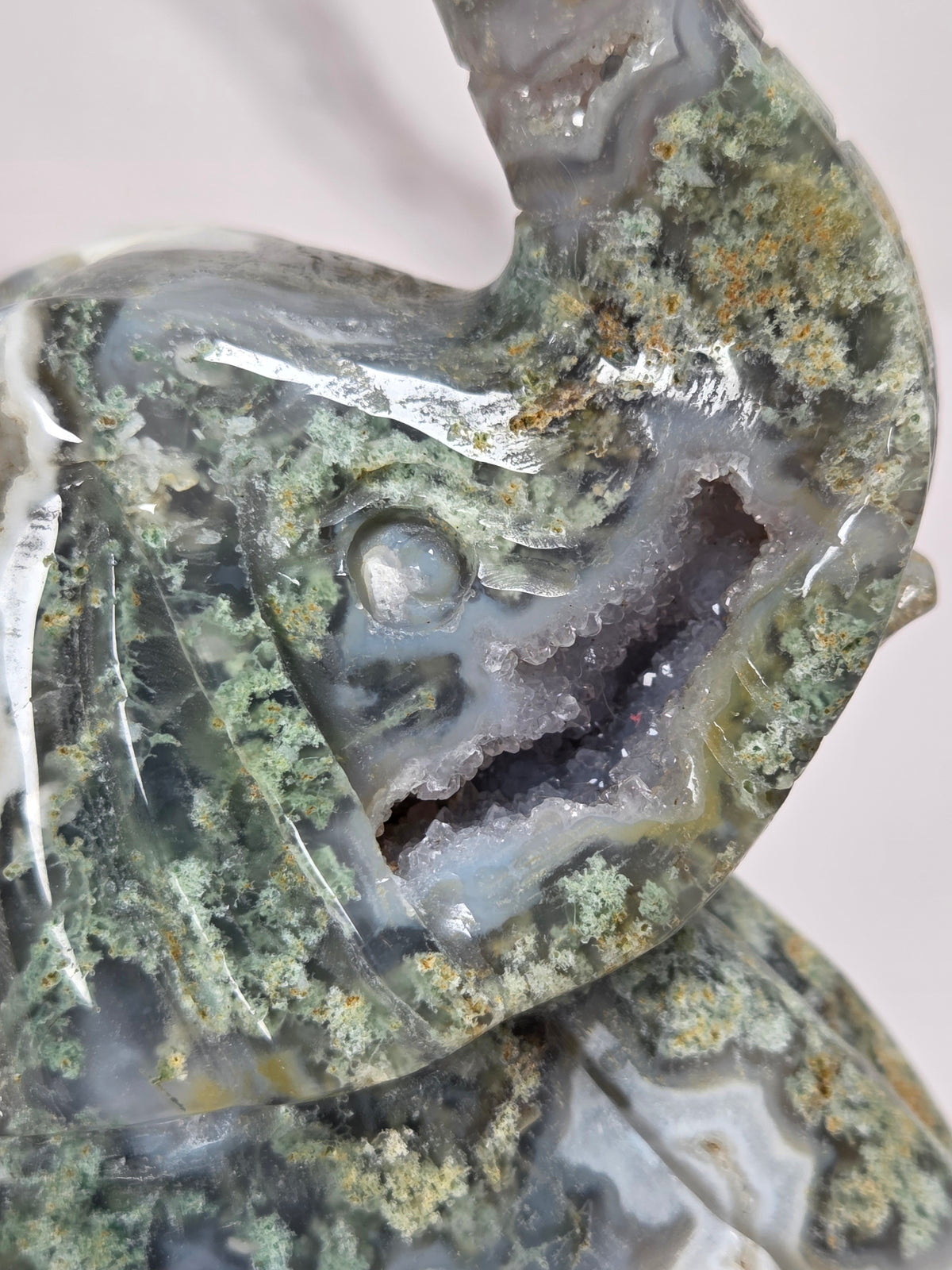 A close-up of a polished agate geode figurine with a dragon head shape, featuring green, gray, and white banded patterns with orange mineral deposits.