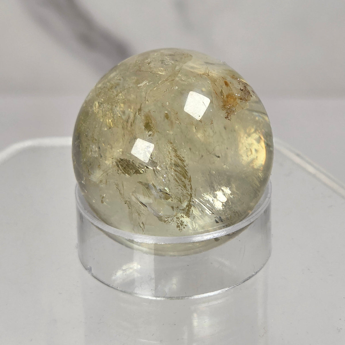A round, polished clear quartz crystal sphere with internal inclusions, displayed on a clear acrylic stand against a light gray marble-patterned background.