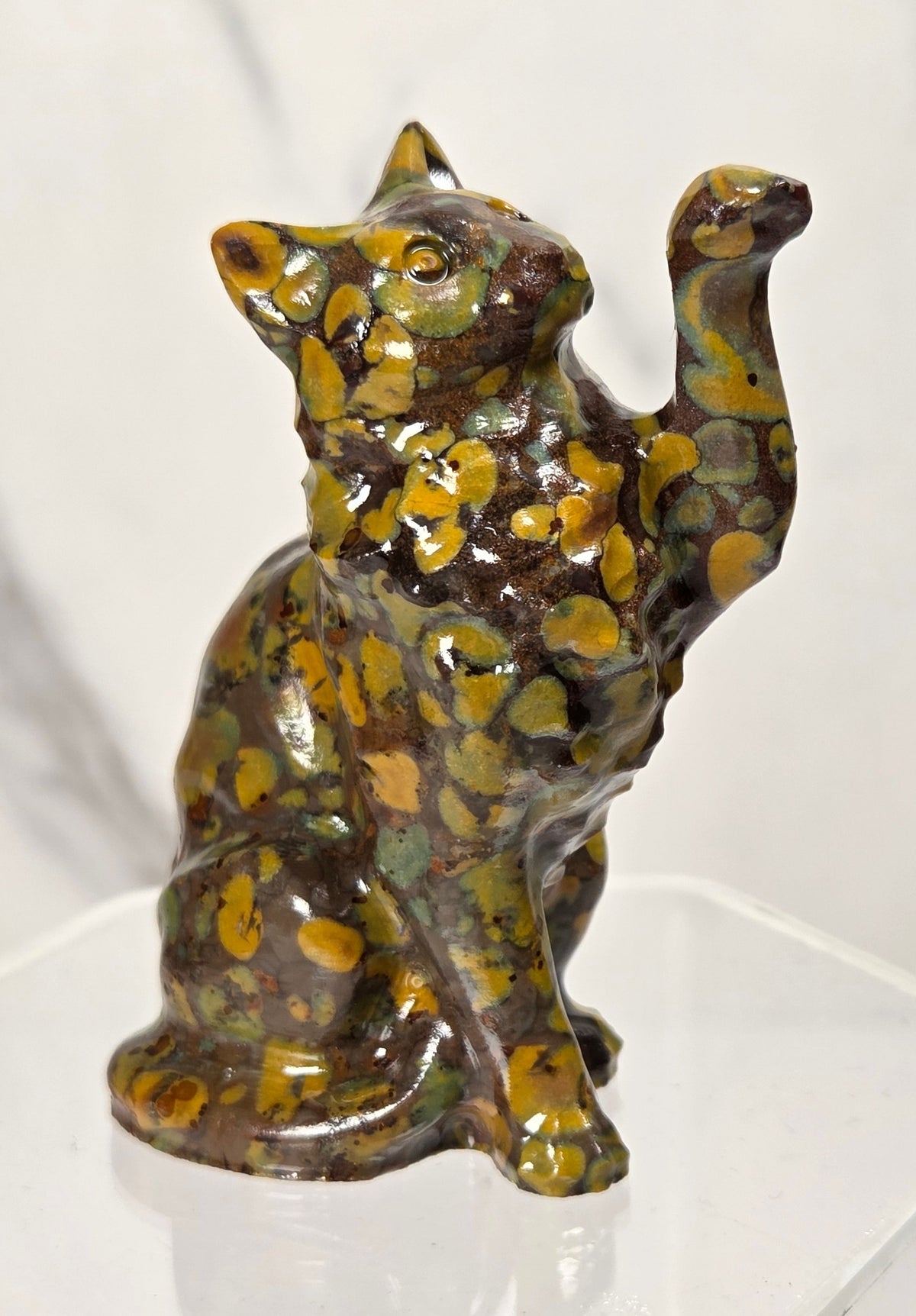 Colorful spotted Ajooba Jasper carving of a cat with it's paw in the air on an acrylic stand against a white background