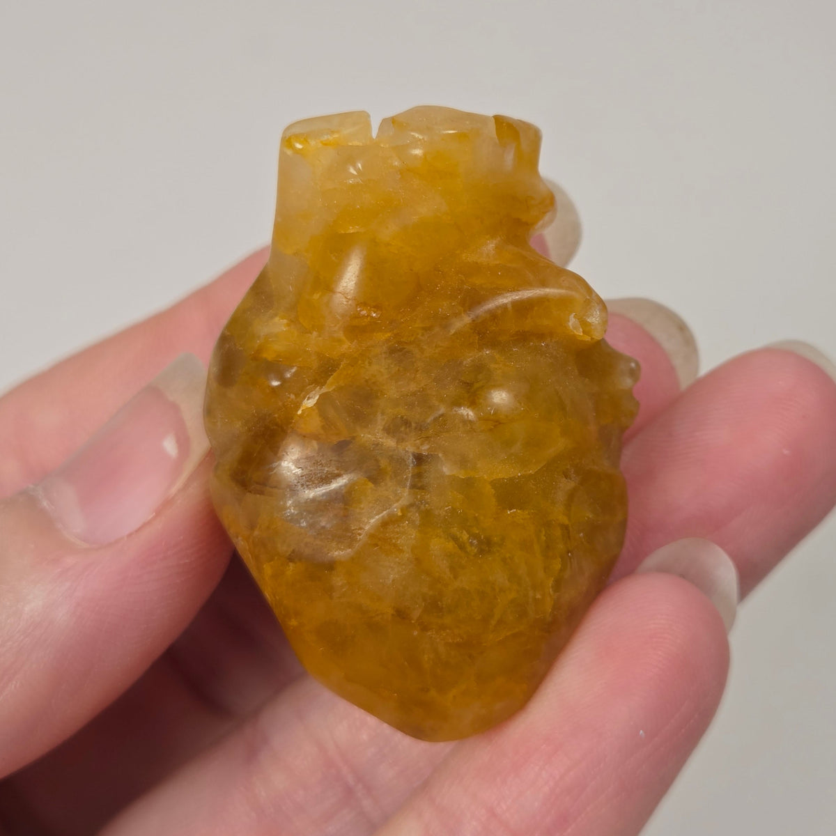 A hand holding a small carved translucent heart-shaped stone pendant with gold coloring, photographed against a softly blurred light background.