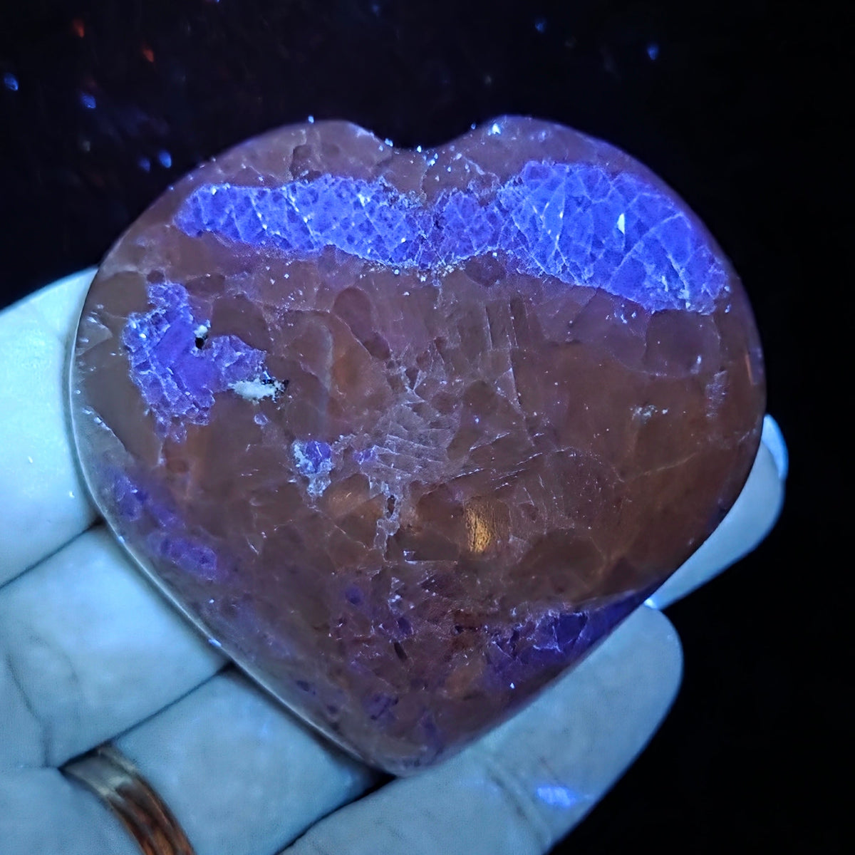 Hand holding a white crystal heart with purple coloring under UV light against a black background