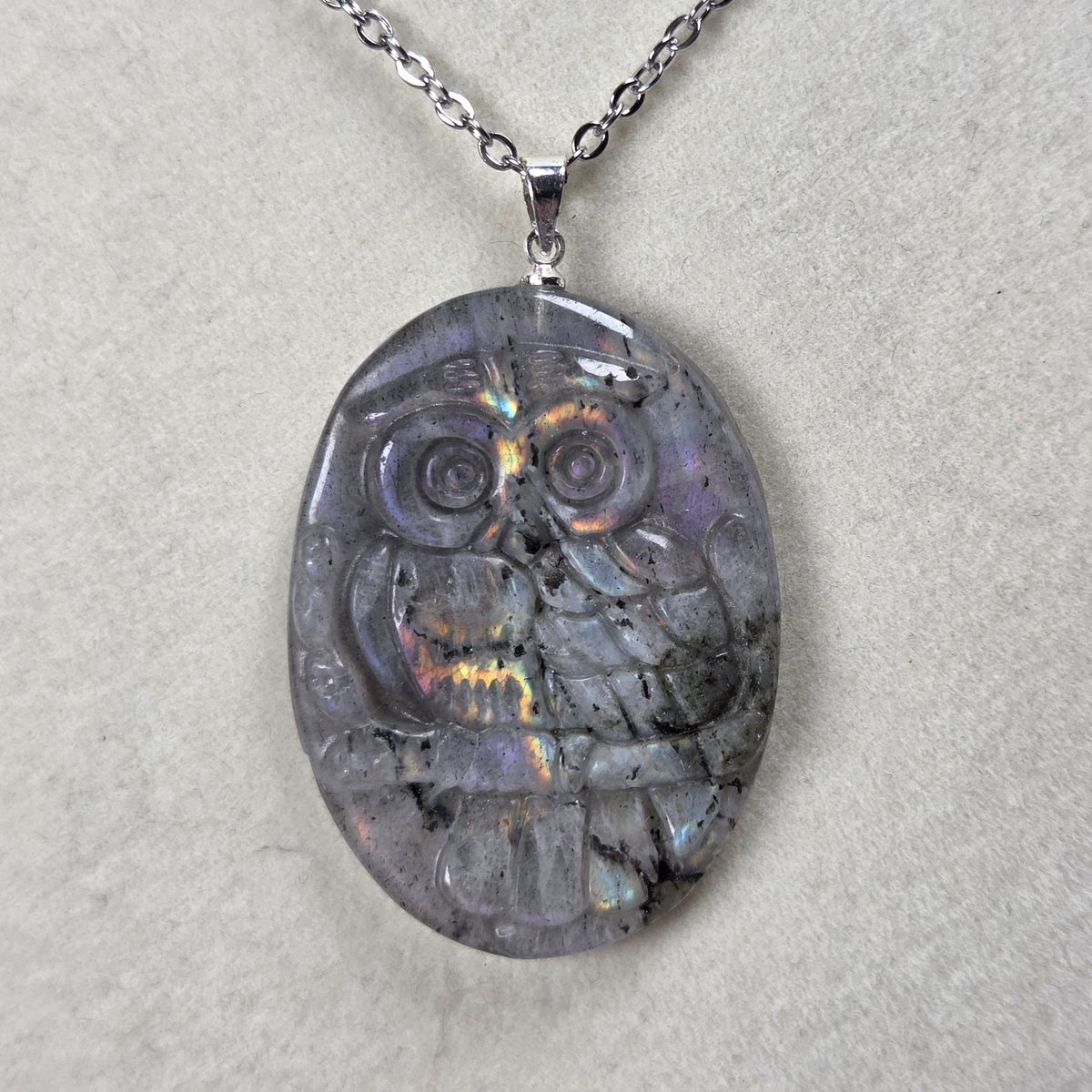 Grey crystal owl shaped pendant with rainbow flash on a silver chain against a tan background