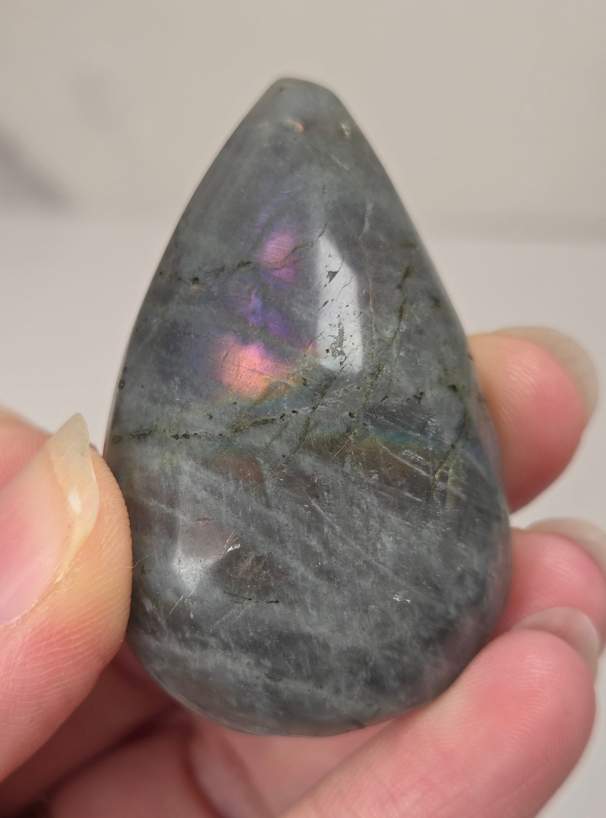 Hand holding a grey labradorite carved raindrop shaped carving with rainbow coloring against a white background