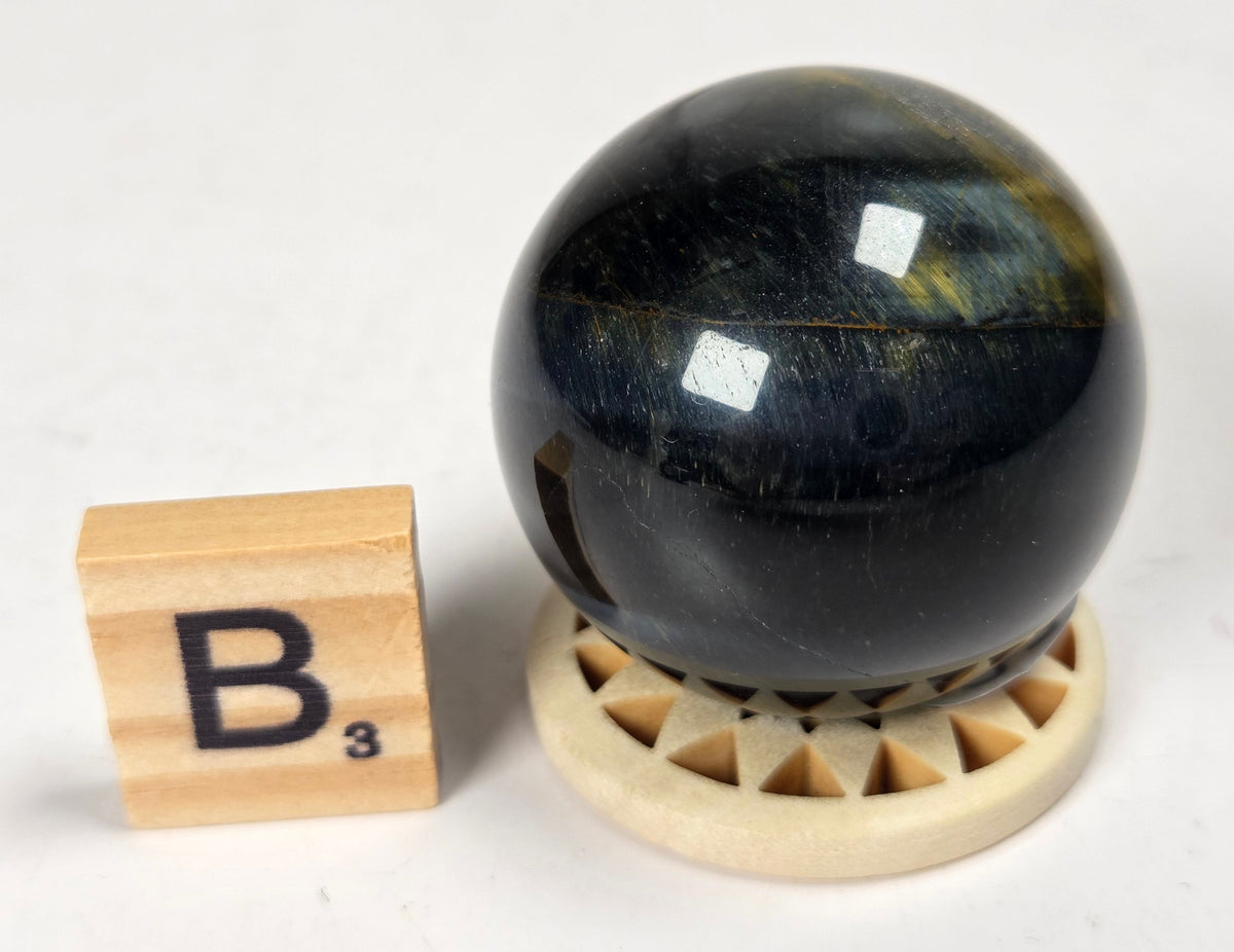 Blue Tigers Eye polished sphere carving with blue and yellow coloring on a wood stand with a scrabble tile against a white background