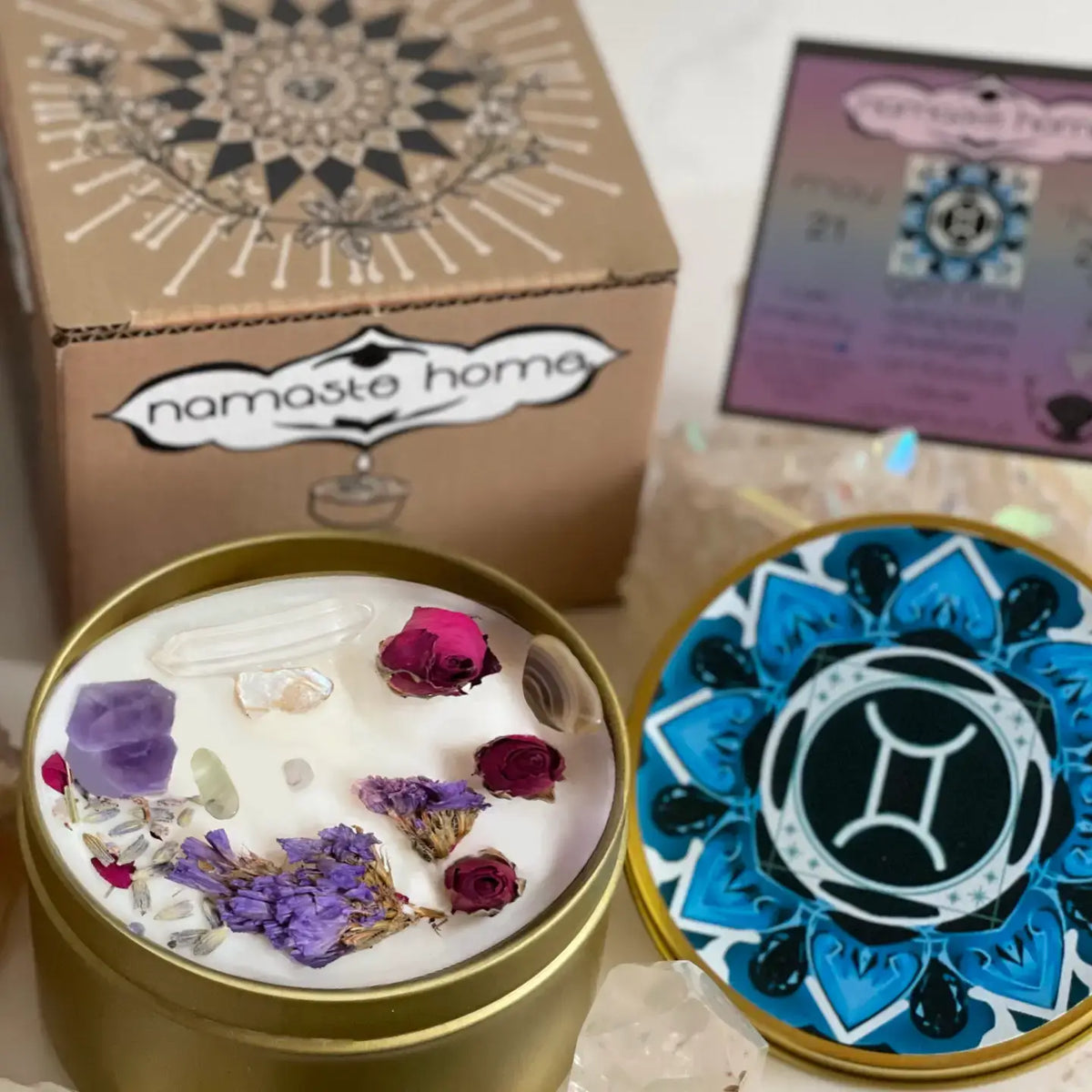 A gold metal candle tin filled with white wax and dried botanicals and crystals, shown beside its blue and black patterned lid and a branded kraft box labeled namaste home.