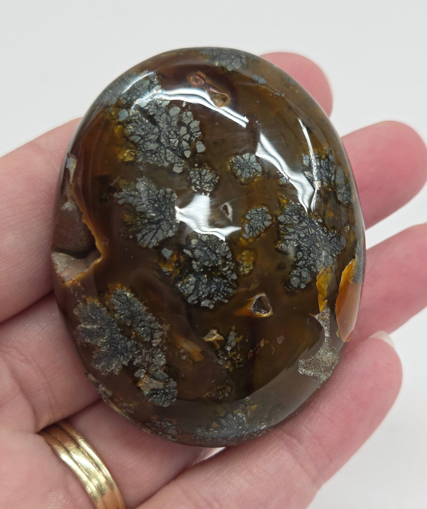 Hand holding a polished marcasite stone with various shades of red, orange, gold, and gray, displayed on a white surface.