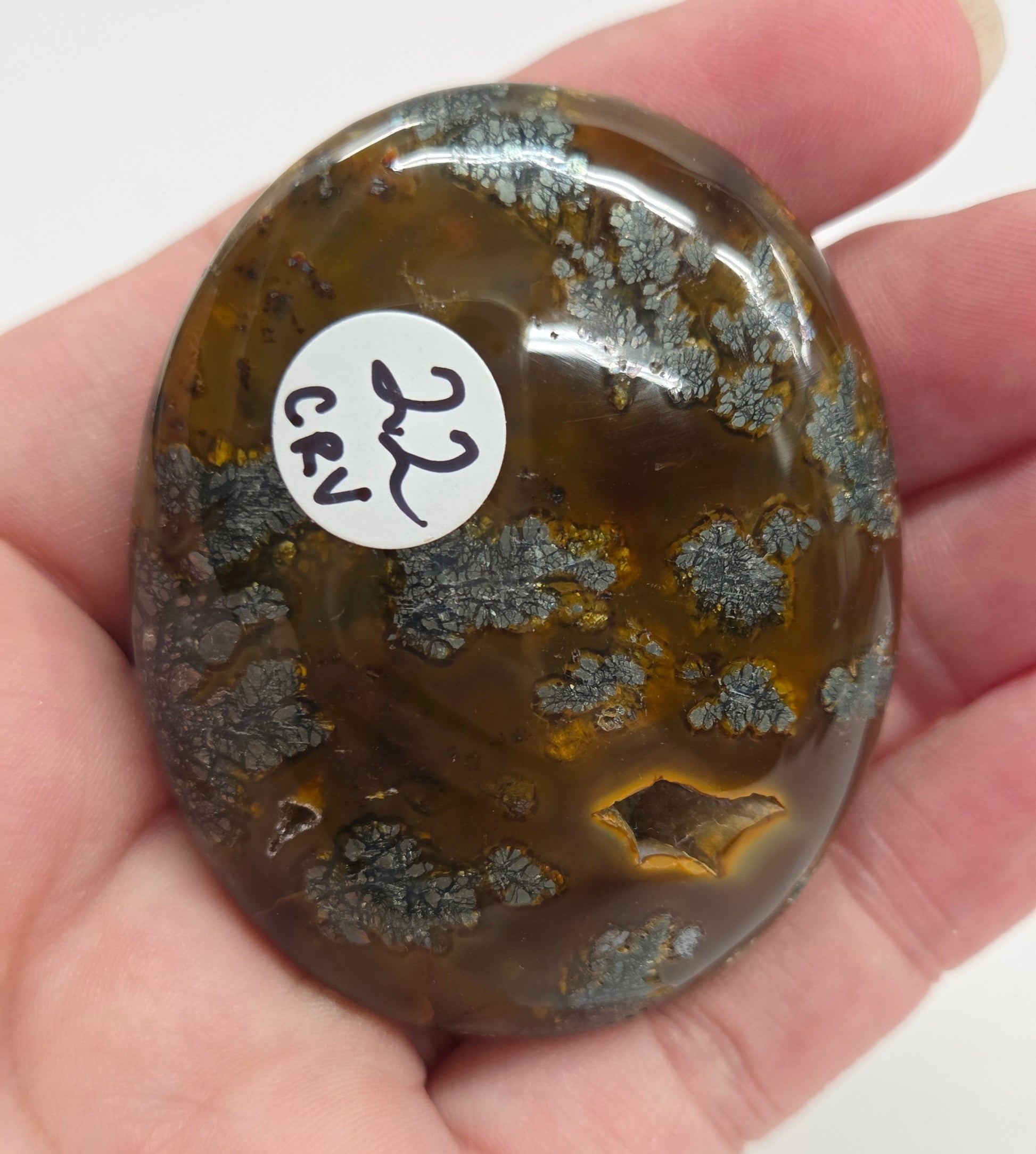 Hand holding a polished marcasite stone with various shades of red, orange, gold, and gray, displayed on a white surface.
