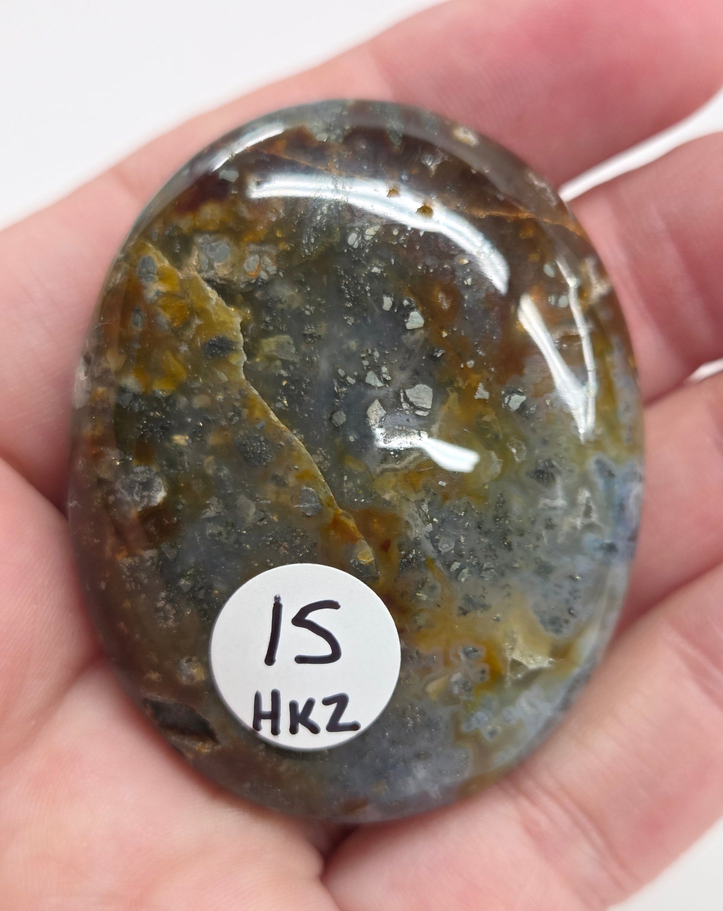 Hand holding a polished marcasite stone with various shades of red, orange, gold, and gray, displayed on a white surface.