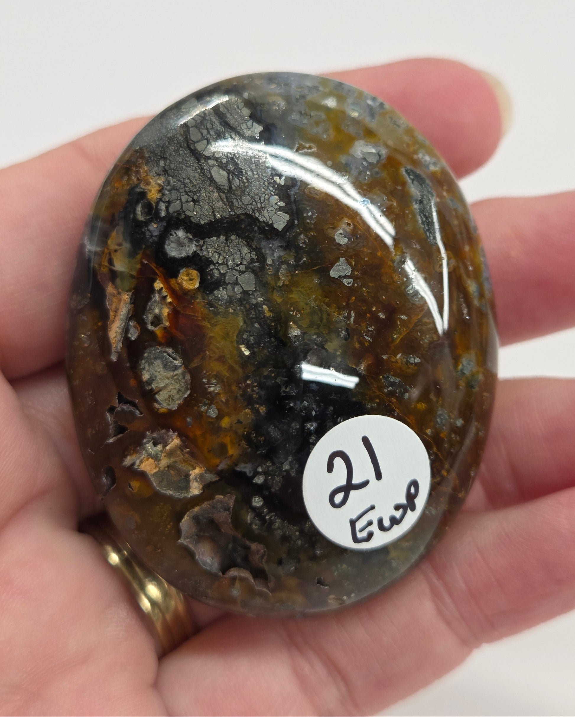 Hand holding a polished marcasite stone with various shades of red, orange, gold, and gray, displayed on a white surface.