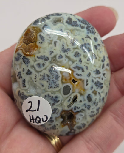 Hand holding a polished marcasite stone with various shades of red, orange, gold, and gray, displayed on a white surface.
