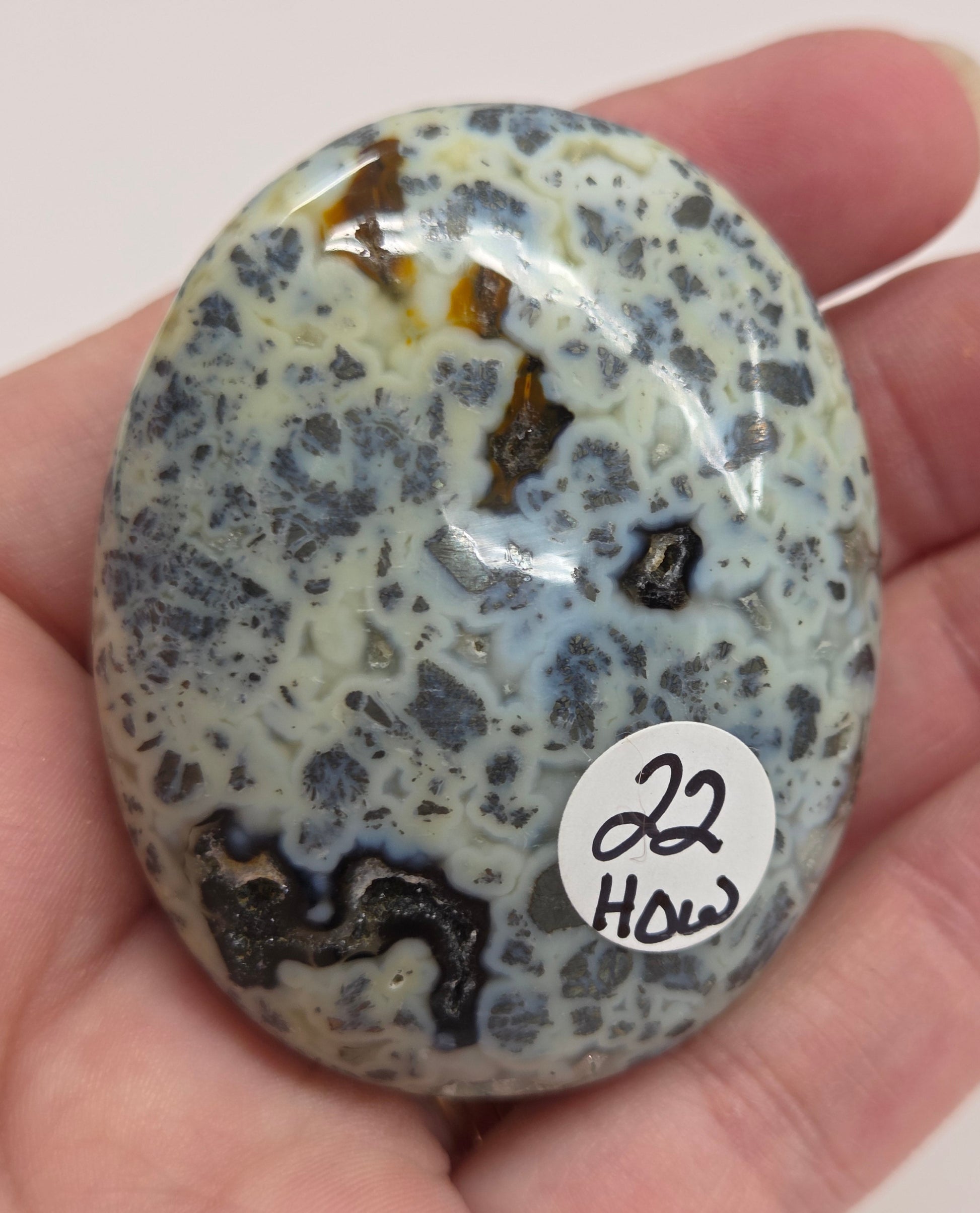 Hand holding a polished marcasite stone with various shades of red, orange, gold, and gray, displayed on a white surface.