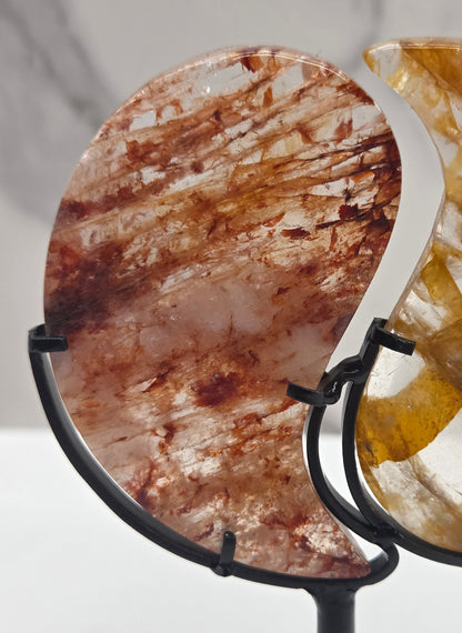 Two stones carved into a yin yang symbol, one fire quartz, one golden healer, on a black stand against a marble background