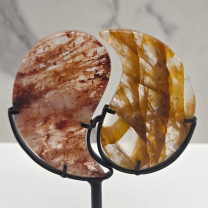 Two stones carved into a yin yang symbol, one fire quartz, one golden healer, on a black stand against a marble background