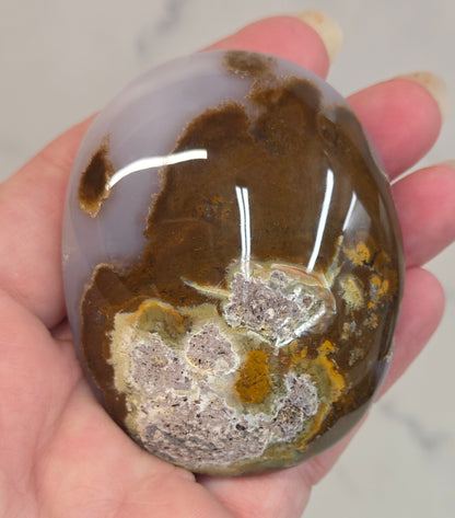 Hand holding a Scenic Moss Agate palm with white, yellow, red and brown coloring on a white background