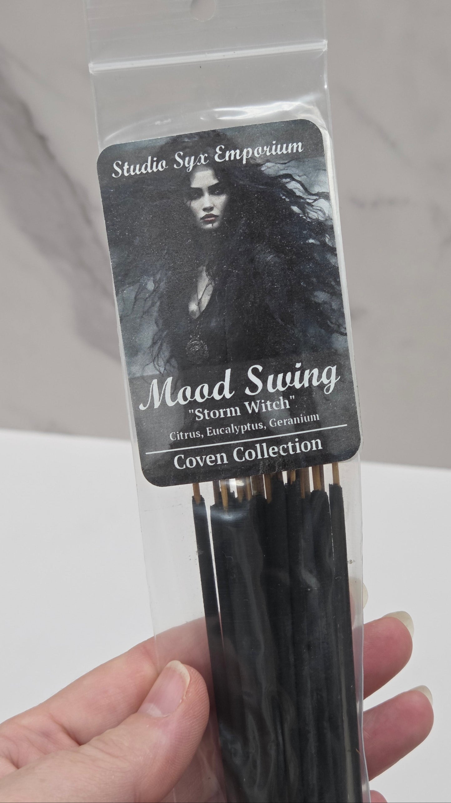 A hand holding a pack of Studio Syx Emporium incense sticks from the Coven Collection, specifically the Mood Swing scent. The packaging is clear and beige, showing the incense sticks inside.