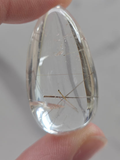 Clear quartz teardrrop shaped crystal with gold rutile inclusions held between fingers