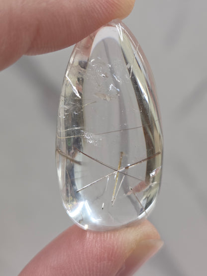 Clear quartz teardrrop shaped crystal with gold rutile inclusions held between fingers