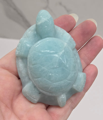 Hand holding a polished Caribbean Calcite crystal in the shape of a sea turtle, with blue and white colors and a light brown pattern.