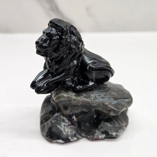 Black Obsidian sculpture of a lion on  a grey yooperlite rock on a white background