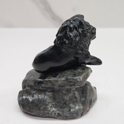 Black Obsidian sculpture of a lion on  a grey yooperlite rock on a white background