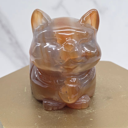 An Agate cat carving, with varying colors of orange, grey and white, on a wood stand against a white background