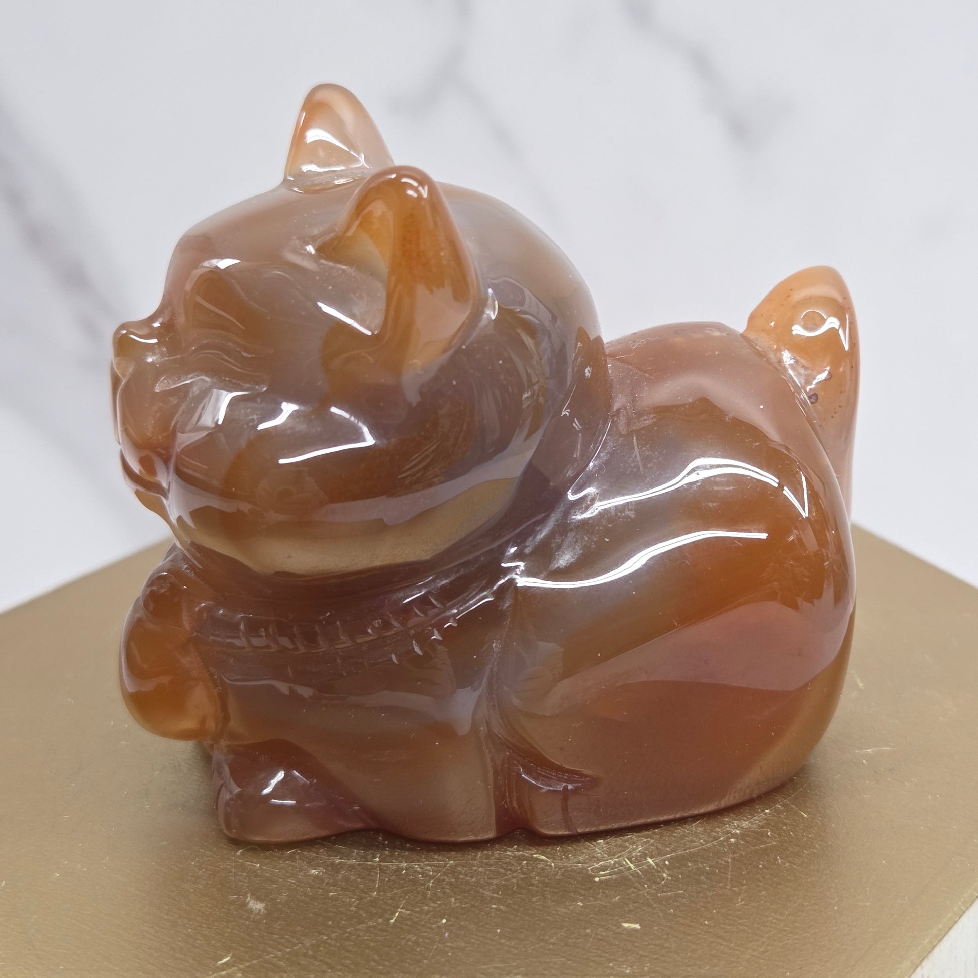 An Agate cat carving, with varying colors of orange, grey and white, on a wood stand against a white background