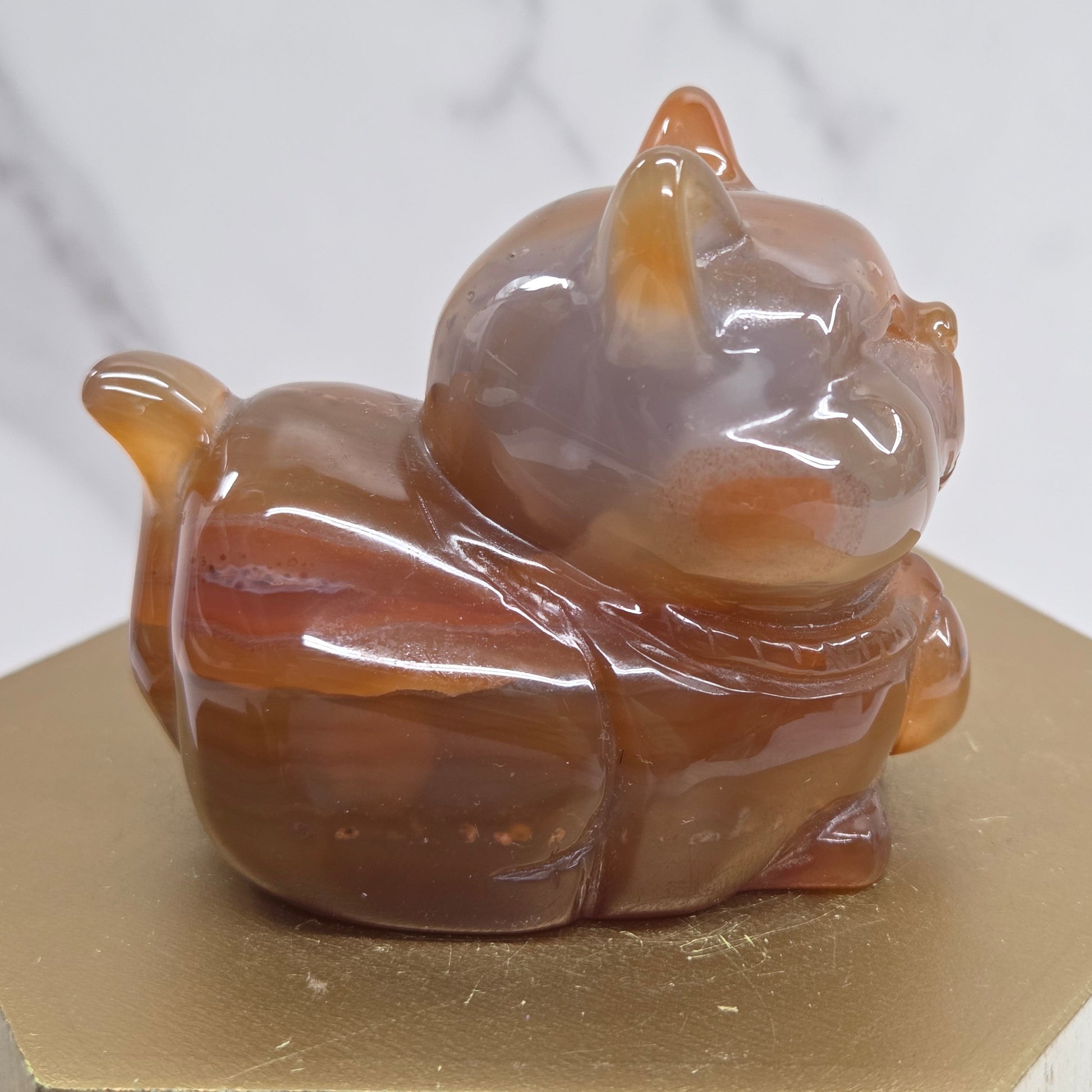 An Agate cat carving, with varying colors of orange, grey and white, on a wood stand against a white background