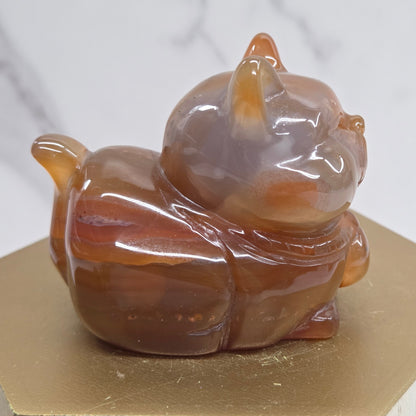 An Agate cat carving, with varying colors of orange, grey and white, on a wood stand against a white background