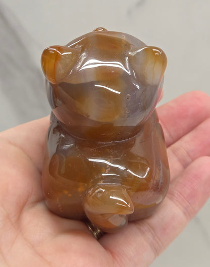 An Agate cat carving, with varying colors of orange, grey and white, being held by a hand against a white background